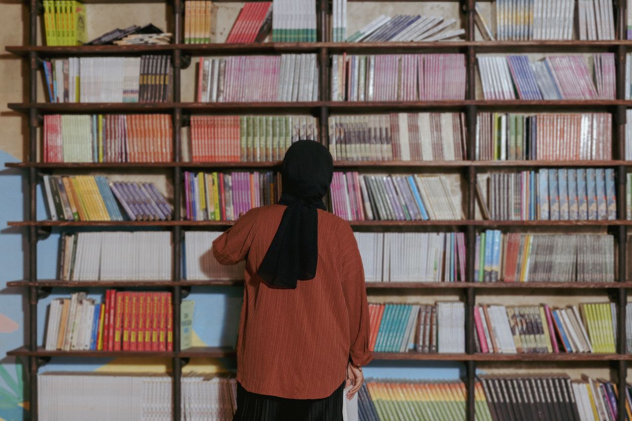 A woman in a headscarf selects a book from a library filled with colorful volumes.