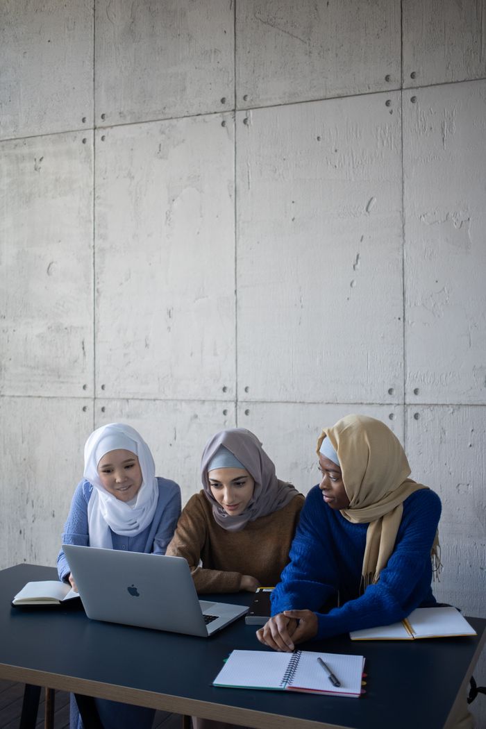 Concentrated multiethnic female students in headscarves sitting at table with copybooks and browsing netbook while doing assignment in classroom together